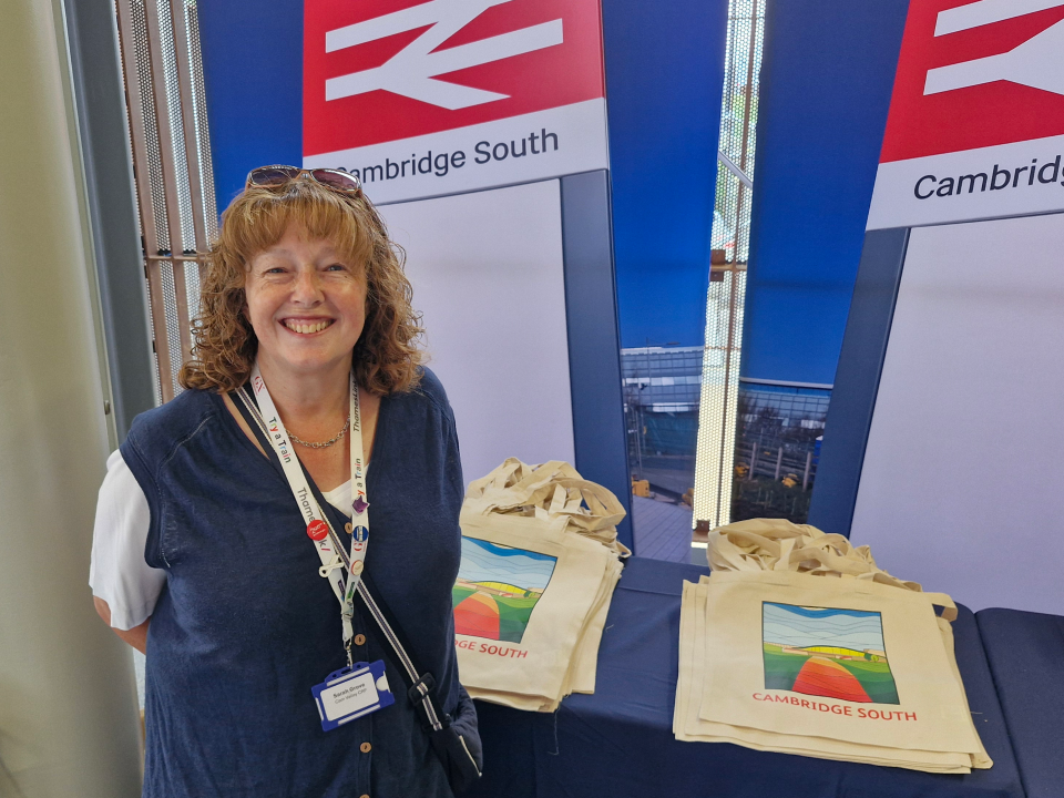 A photograph of Sarah Grove, Project Officer for the Cam Valley CRP. Behind Sarah are tote bags featuring a graphic design of Cambridge South Station.