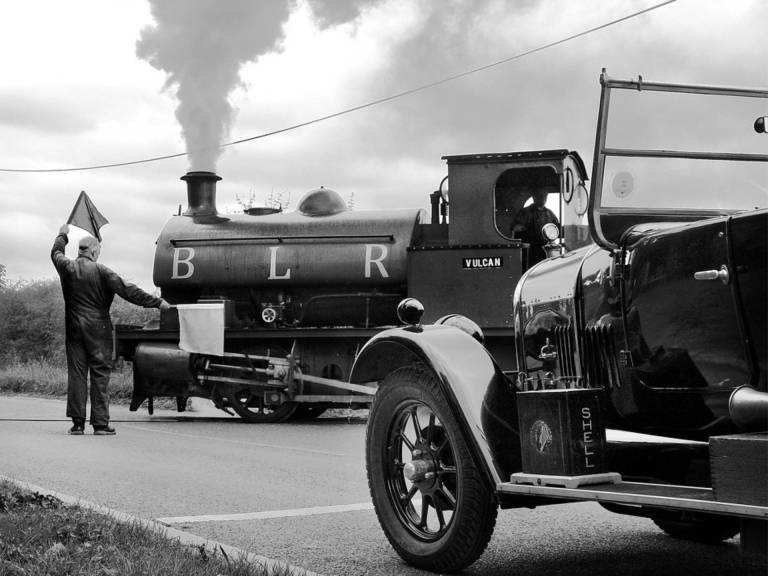 Black and white photograph of a man holding up a car at a railway crossing with flags as a steam train crosses the road.