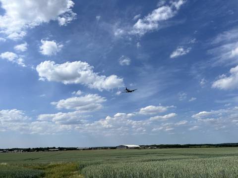 A photograph of Duxford airfield from a distance. The sky is blue with fluffy clouds and a vintage aircraft is flying.
